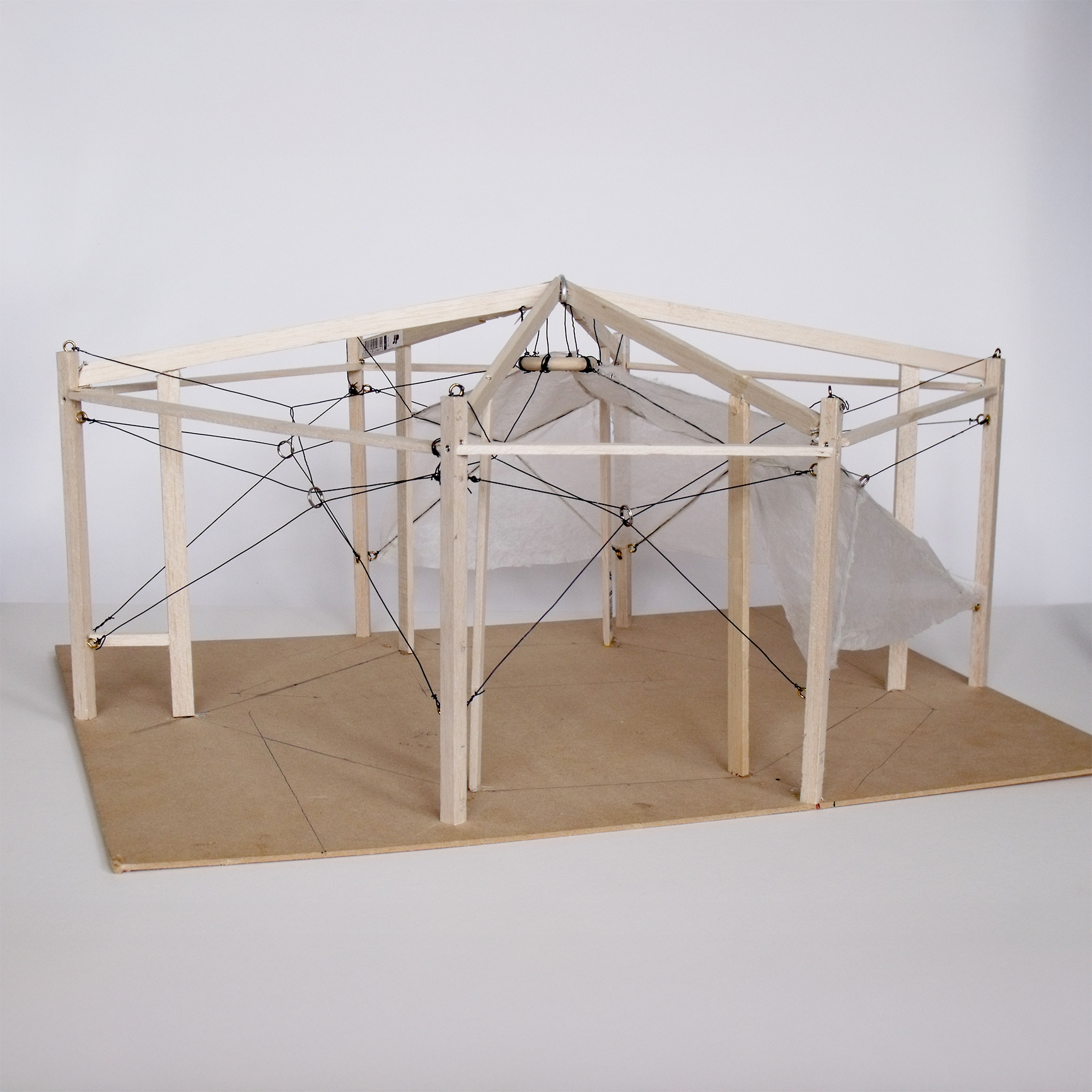 Architectural model made of wooden sticks and black cords forming a geometric structure, with translucent fabric panels suspended inside, placed on a wooden base against a neutral background